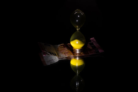 An hourglass and ten euros lie on a mirror in the dark, time is money, business and finance