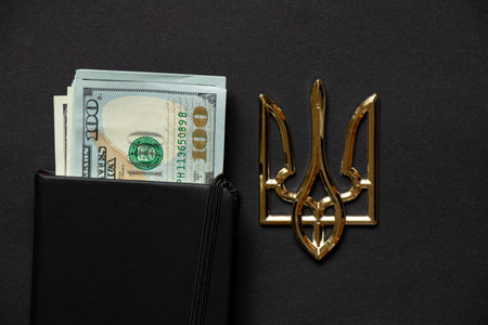 One hundred dollars lie in a white blank notepad on black and next to it lies the coat of arms of Ukraine, business and income in Ukraine, a notepad and space for textの写真素材