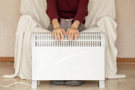 A girl sits on an armchair and puts her hands on a warm convector in an apartment in winter, a heating season, a warm house in Ukraineの写真素材