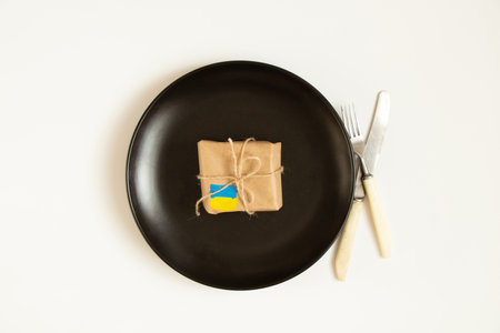 A box with a painted flag of Ukraine lies on a black plate and next to a knife with a fork on a white background, humanitarian aid for Ukraine, warの写真素材