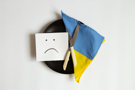 A sad emoticon is drawn on paper, lies on a plate next to the flag of Ukraine on a white background,...