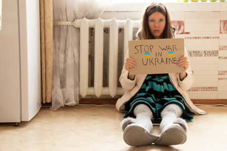 A girl sits in a winter jacket sits near a cold radiator in an apartment with a sign stop war in...