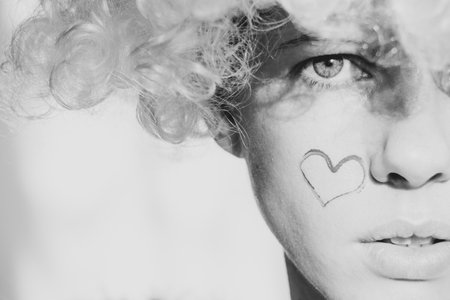Girl dressed as an angel with wings and curly hair with a painted heart on her cheek on an isolated background, an angel and loveの写真素材