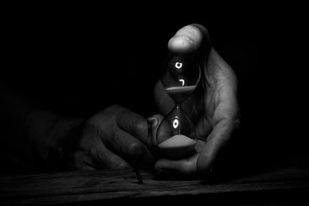 An old female hand holds an hourglass in the dark at the table, time goes by, the speed of life
