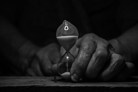 An old female hand holds an hourglass in the dark at the table, time goes by, the speed of life
