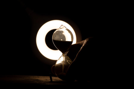 An old female hand holds an hourglass in the dark at the table, time goes by, the speed of life