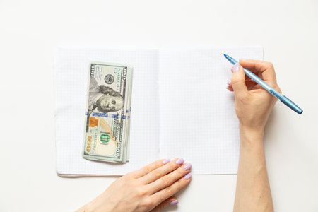 Women's hands write in a notebook, and dollars lie nearby on a white background, businessの写真素材