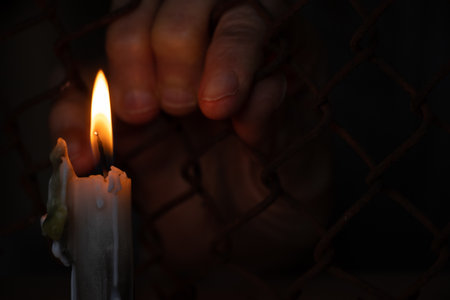 old female hands hang on a fence made of iron mesh in the lock on the background of a burning candleの写真素材