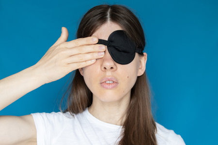 A girl with a bandage on one eye and the other eye closed with her hand on a blue background, don't see, don't lookの写真素材