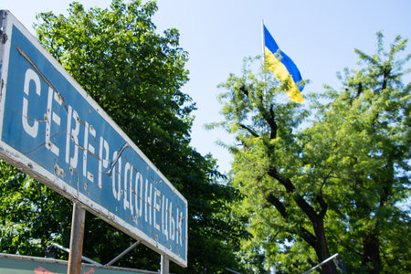 The name of the city in Ukrainian Severodonetsk on a road sign broken from bullets against the background of the flag of Ukraine in the sky, a city in Ukraineの写真素材