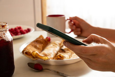 A woman holds a cup of coffee and a phone and has breakfast with pancakes with raspberries in the kitchen at home, working online in the phone, ordering foodの写真素材