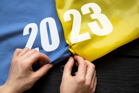 Women's hands sew the flag of Ukraine on which the text 2023 at the table, work and patriotism