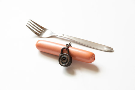 Sausage on a chain under lock and key and a fork next to it on a white background in the sun, rejection of unhealthy food, dietの写真素材