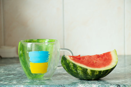 A cup with a painted flag of Ukraine and a sliced watermelon next to it from the city of Kherson in Ukraine in the kitchen of a house in Ukraineの写真素材