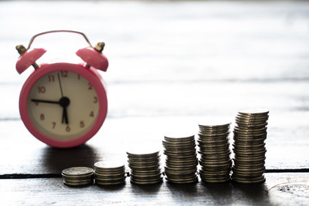 Stacks of coins stand on a wooden table and an alarm clock stands nearby, business and finance, success timeの写真素材