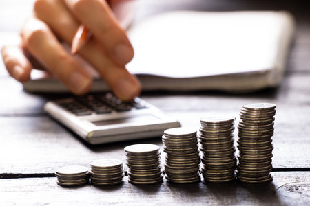 A hand on a calculator and next to it lies a notebook and stacks of coins, business and financeの写真素材