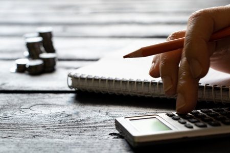 A hand on a calculator and next to it lies a notebook and stacks of coins, business and financeの写真素材