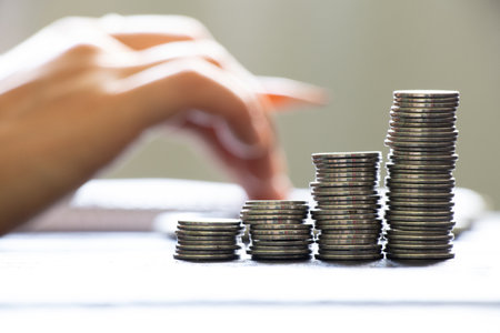 A hand on a calculator and next to it lies a notebook and stacks of coins, business and financeの写真素材