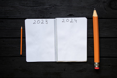 The year 2023 and 2024 is written on the pages of a notebook and next to it lies a large and small pencil on a black table, the end of the old year and the beginning of a new oneの写真素材