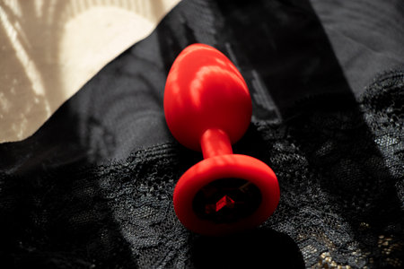 A red silicone smooth butt plug, a sex toy, lies on black lace panties in the shade. Contrast photo of anal toys, erotic toysの写真素材