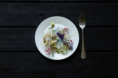 White plate with various dried flowers on a black wooden board and a fork nearby, flowers and foodの写真素材