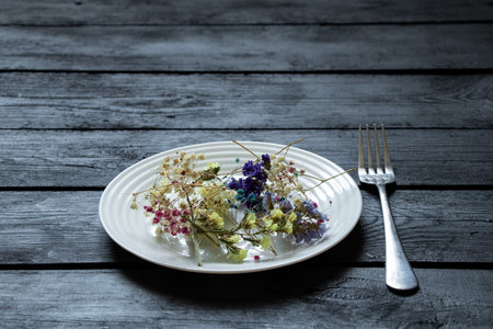 White plate with various dried flowers on a black wooden board and a fork nearby, flowers and foodの写真素材