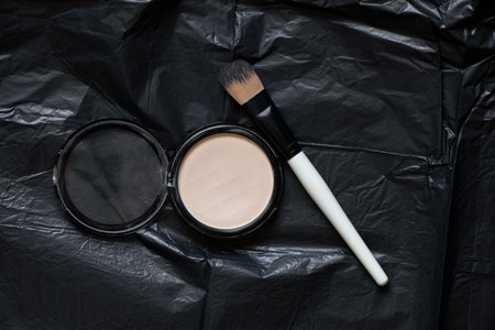 Plastic face foundation and powder brush lie on crumpled black paper, cosmetics
