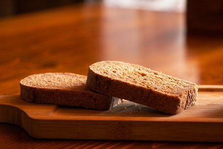 Two pieces of sliced black bread lie on a wooden board on a table in the sun, bread on the table