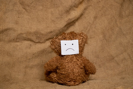 A brown bear sits with his back, and on his back there is a paper drawn with a sad face, against the background of burlap, sadnessの写真素材