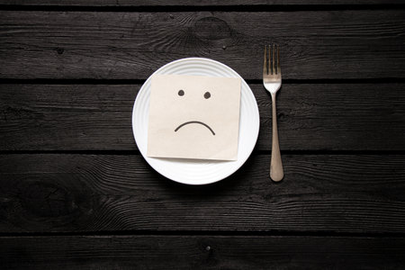 A white plate and paper with a drawn breast stand on a black wooden table and a fork next to it, nothing to eat, hungerの写真素材