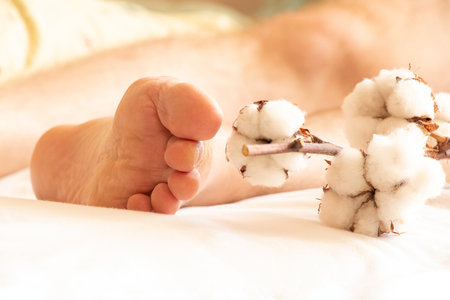 Male leg lies on bed near dry cotton branch at home, male bare foot, beauty and healthの写真素材