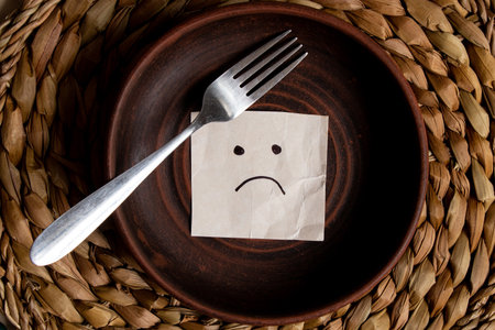 A card with sadness drawn on it lies on an empty plate on a straw background and next to it is a fork, no food, hunger and nothing to eatの写真素材