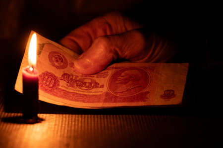 An old womans hand holds a 10 ruble note from 1961 near a candle flame in the dark, ten rubles USSR