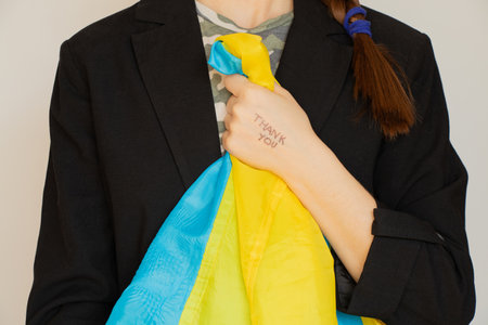 Thank you is written on the hand of a girl holding a Ukrainian flag and wearing a black business jacket and a camouflage T-shirt, thank youの写真素材