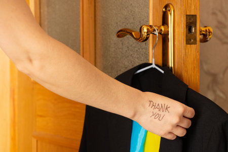 Girl with written text thank you on her hands holds business black jacket and Ukrainian flag on hanger at home, words of gratitudeの写真素材