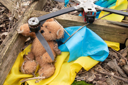 A downed FPV drone lies amidst the ruins of a destroyed house, alongside a child's teddy bear and a flag in Ukraineの写真素材