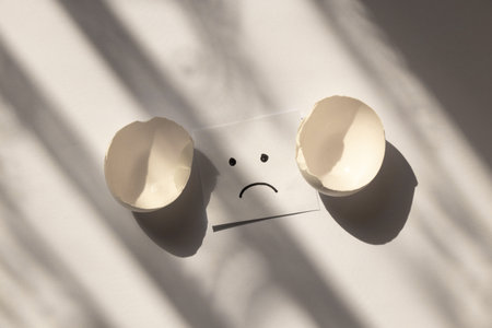 Chicken egg shell broken and sad emotions drawn on paper on white background in shadowの写真素材