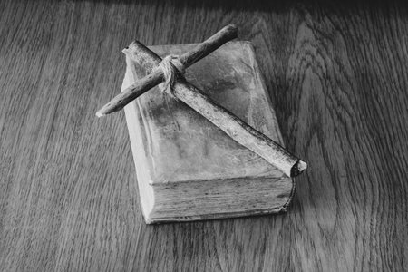 A wooden homemade cross made of branches tied with a rope lies on a book on a wooden table, prays, faith in God and religionの写真素材