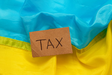 The inscription tax on a piece of cardboard against the background of the blue and yellow flag of Ukraine, the concept of taxes in Ukraine, economicsの写真素材