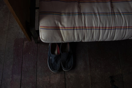 Old men's slippers stand on a wooden floor near a bed in a house without renovation, shoes near the bedの写真素材