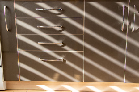 Kitchen brown drawers with handles in the apartment on the kitchen with a striped shadow from the curtains on the windowの写真素材