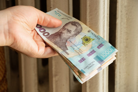 1000 UAH in the hands of a man near a white radiator in an apartment in Ukraine, tariff for central heating in an apartmentの写真素材