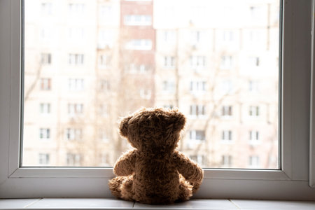 A teddy bear sits on a windowsill in an apartment and looks out the window at a multi-story building in Ukraineの写真素材