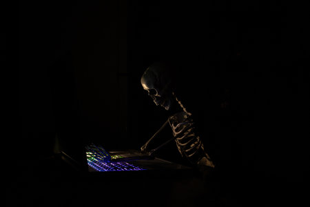Skeleton working on laptop with backlit keyboard in dark at home, skeleton near laptopの写真素材