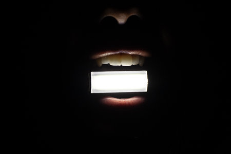 The girl holds a small flashlight in her teeth in complete darkness, shines a flashlight, without lightの写真素材