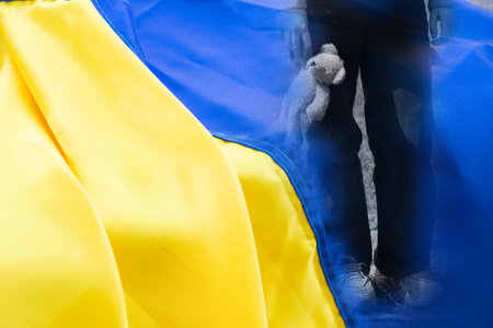 Little girl with a teddy bear in her hand on the background of the Ukrainian flagの写真素材