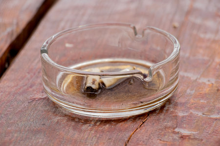 Cigarette butts in a glass ashtray with water on a wooden surface, smoking and nicotine addictionの写真素材