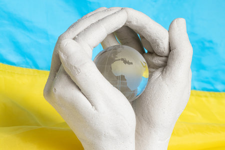 Plaster hands holding glass globe against Ukrainian flag background, concept of peace, protection and responsibility for the planetの写真素材