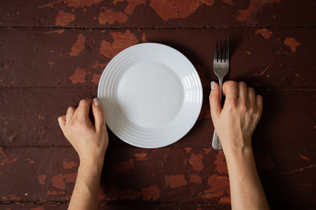 Female hands with fork and empty plate on old wooden table, concept of hunger, waiting for food, diet, poverty or fastingの写真素材