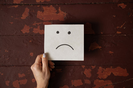 Female hand holding a card with a sad face drawn on the background of an old wooden brown table, emotion of sadness and discontentの写真素材